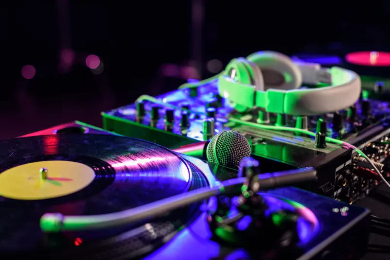 Nasmejana DJ-ka drži retro vinilno ploščo, medtem ko stoji za svojo DJ opremo v zadimljenem nočnem klubu, pripravljena za mixanje in zabavo.
