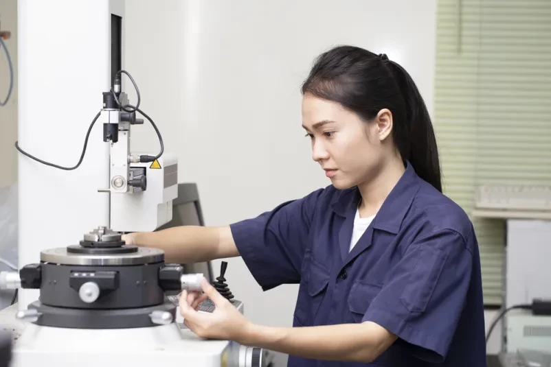 Mlada inženirka azijskega porekla v industrijskem laboratoriju testira in nastavlja kompleksno strojno opremo, kar kaže na zagotavljanje skladnosti izdelkov.
