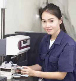 Inženirka v modri uniformi nastavlja sodoben industrijski stroj v laboratoriju, kar je del procesa zagotavljanja kakovosti v livarnah.