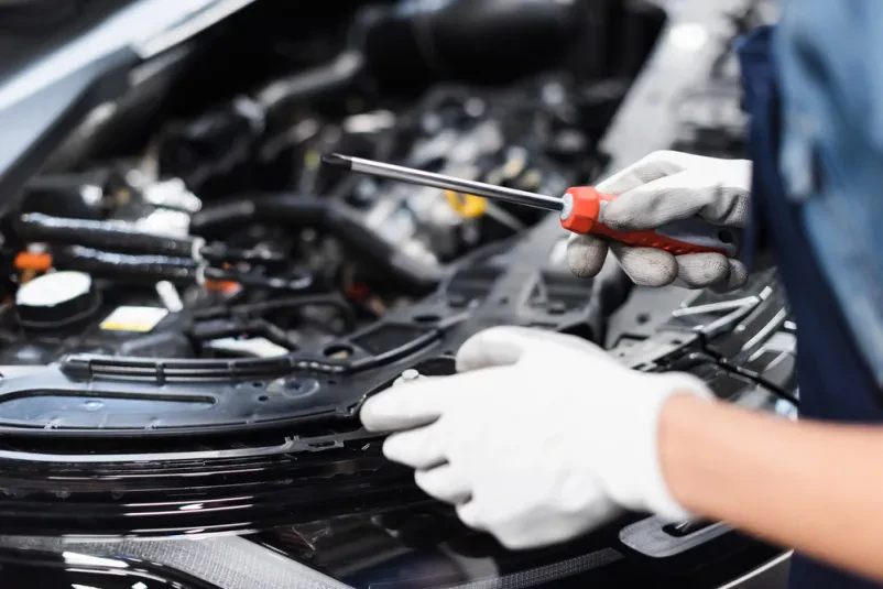 Detailni posnetek ženske mehanik v zaščitnih rokavicah, ki s križnim izvijačem natančno popravlja avtomobilski motor v garaži.