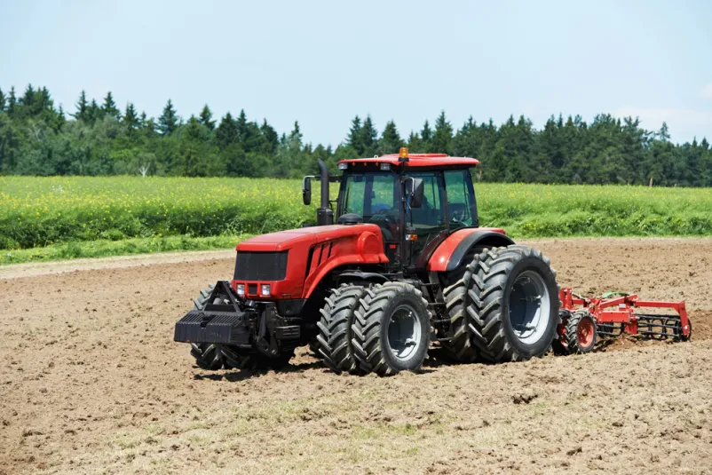 Masiven traktor z robustnim plugom temeljito obdeluje polje, dvigajoč svežo prst. Priprava tal je ključno kmetijsko delo za uspešno sezono.