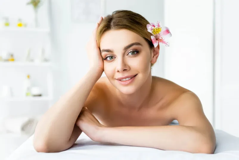 Sproščena ženska na masažni mizi s cvetom Nasmejana ženska sproščeno leži na masažni mizi, z belim cvetom na hrbtu. Prizor nakazuje globoko sprostitev in dobro počutje med wellness tretmajem.