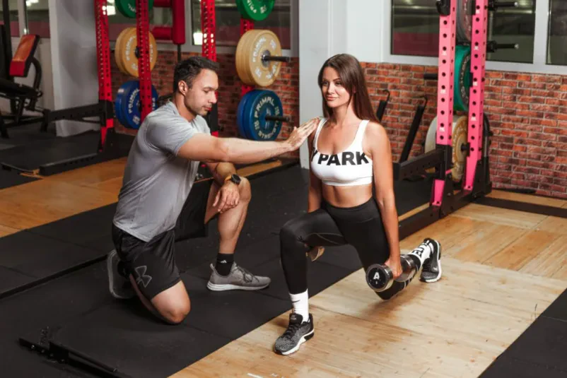 Profesionalni osebni trener v fitnesu Maribor pomaga stranki pri pravilni tehniki dvigovanja uteži v dobro opremljenem fitnes centru.