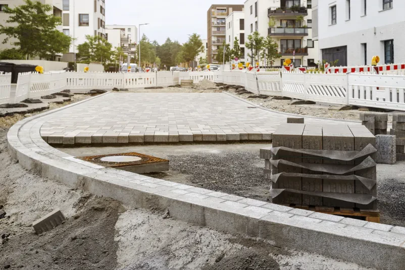 Prikaz del na gradbišču, kjer se polagajo tlakovci za prenovo cestne površine. Delo poteka ob zaobljenem modernem odseku ceste, kar kaže na strokovno tlakovanje poti.