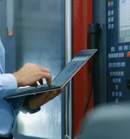 Starejši inženir z vizirjem pred nadzorno ploščo CNC stroja, ki programira nastavitve. Poudarjena je kompleksna programska in krmilna tehnika.