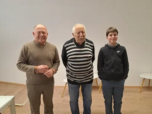 Odrasli udeleženci turnirja so osredotočeni na strateško igro. Prikazano je strastno učenje šaha Idrija, okolica, ki povezuje ljubitelje vseh generacij.