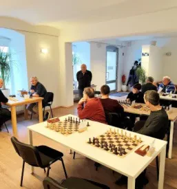 Dva moška igrata šah, eden izmed njiju se nasmiha, kar prikazuje sproščeno vzdušje in prijetno druženje ob šahovski deski, ki poteka med prijatelji.