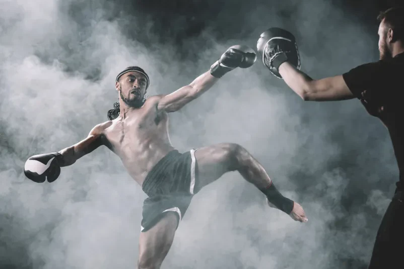 Muay Thai borec izvaja visok udarec Osredotočen Muay Thai borec, izoliran na črnem ozadju, izvaja močan udarec z nogo. Prikazuje intenzivnost tega športa.