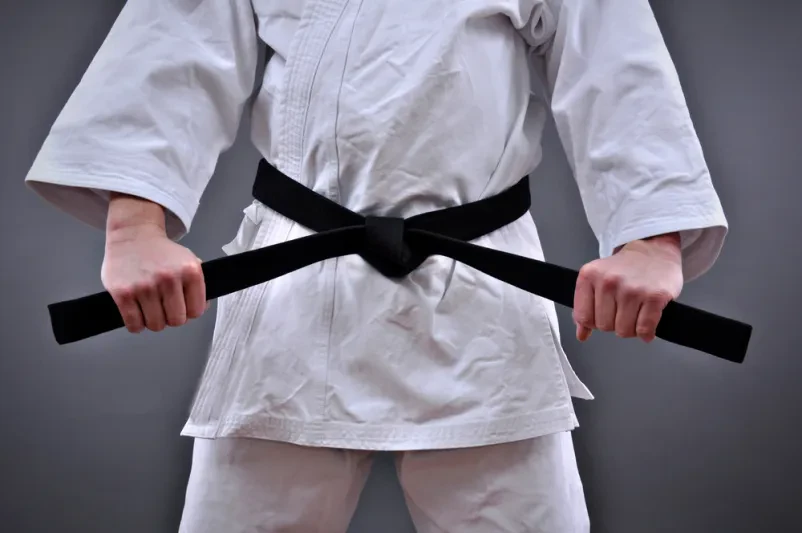 Moški drži svoj črni pas borilnih veščin Moški v uniformi borilnih veščin ponosno drži svoj črni pas z obema rokama, simbol discipline in dolgoletnih dosežkov.