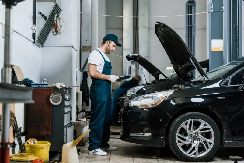 Mehanik s kapo in brado drži pisalo in beležko ob avtu, pripravljen za Castrol avtoservis in pregled vozila.