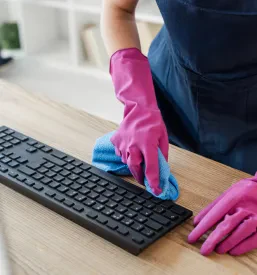 Približan pogled na roko čistilca v gumijasti rokavici, ki s krpo skrbno čisti računalniško tipkovnico v pisarni, kar poudarja natančnost pri čiščenju elektronske opreme.