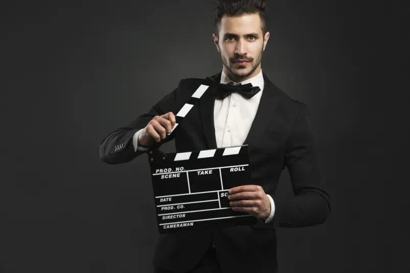 Portret elegantnega moškega v smokingu, ki drži filmsko klapo. Slika poudarja profesionalnost v filmski industriji.