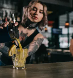 Tetovirani bartender s spretnimi gibi pripravlja koktajl za pultom, obkrožen z različnimi alkoholnimi in brezalkoholnimi pijačami.