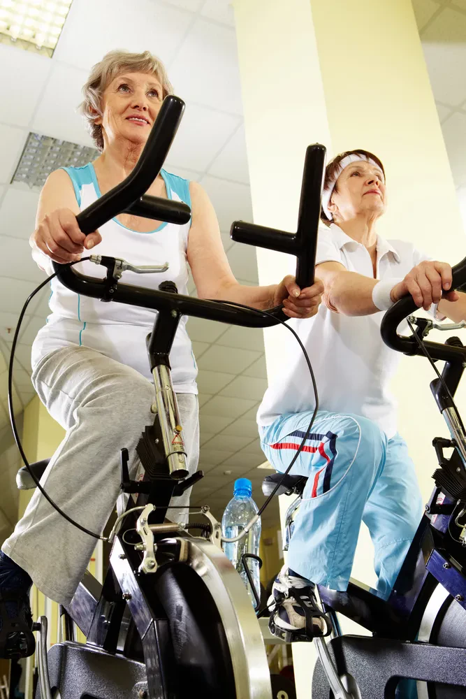 Portret starejše ženske, ki v fitnes klubu z nasmehom izvaja telesno vadbo na posebni opremi in ohranja aktivnost.