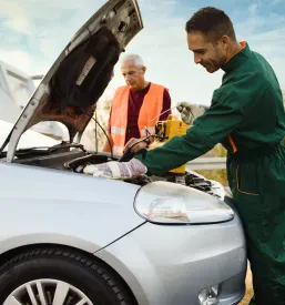 Dva delavca cestne asistence s pomočjo zagonskih kablov poskušata zagnati motor avtomobila, kar je pogosta storitev popravila in vzdrževanja vozil.