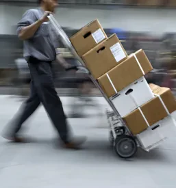 Delavec potiska ročni voziček z naloženimi paketi, kar poudarja učinkovito in hitro dostavo paketov do prejemnika.