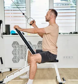 Moški v športnih oblačilih vadi na napravi za moč v kineziološkem centru. Cilj je povečanje moči in izboljšanje vzdržljivosti.