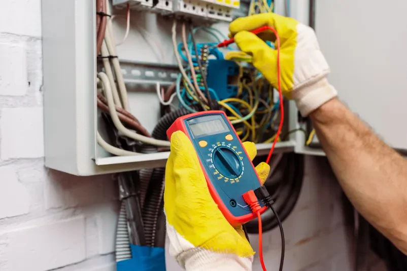 Delavec v rokavicah z multimetrom natančno meri napetost v električnem panelu, zagotavljajoč varnost in delovanje inštalacij.