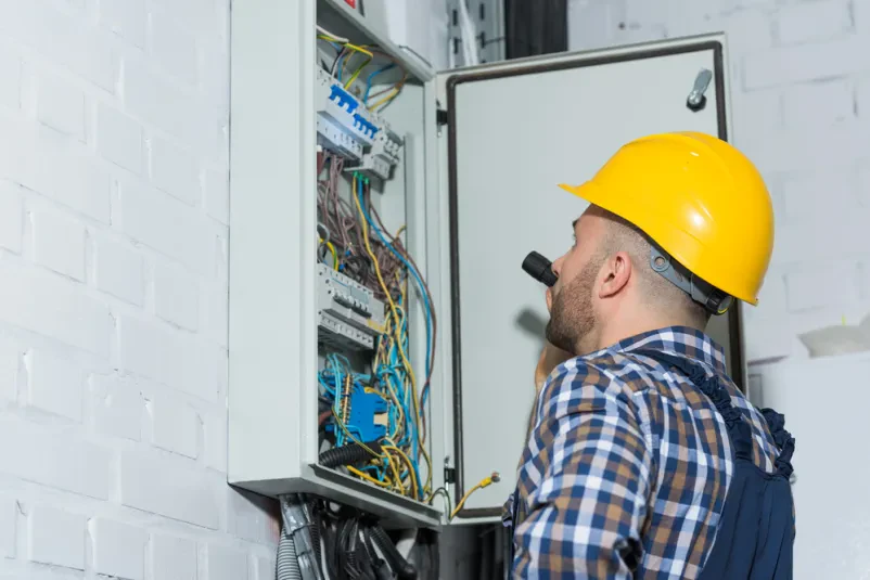 Izkušen električar s svetilko skrbno pregleduje in popravlja razdelilno omarico, kar je ključno za Kvalitetne elektroinštaalcije Hajdina, Podravje.