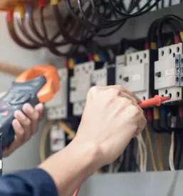 Inženir električar z merilnikom preverja napetost in tok v krmilni omari, s poudarkom na pregledu električnih inštalacij.