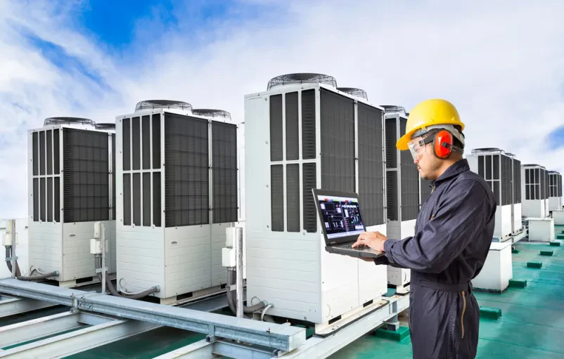 Inženir s prenosnim računalnikom pregleduje industrijski klimatski sistem na strehi tovarne, optimizira delovanje.