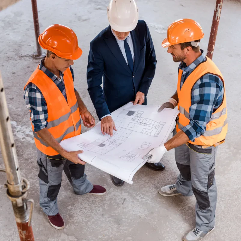Poslovnež in dva gradbenika v čeladah skupaj pregledujejo podrobne modre tlorise z višjega kota, kar poudarja timsko delo na projektu.