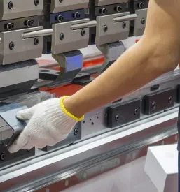 Delavec z varnostnimi rokavicami spretno upravlja stroj za upogibanje, natančno oblikuje pločevinaste dele za nadaljnjo uporabo.