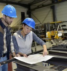 Skupina inženirjev v zaščitnih čeladah razpravlja o načrtih in izzivih v industrijski delavnici, poudarjajoč sodelovanje in strokovnost.
