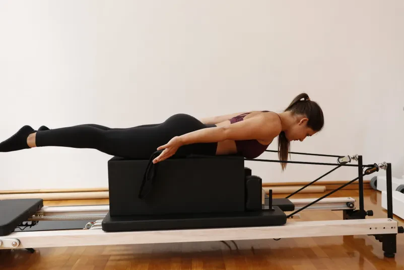 Napredna pilates vaja na reformerju Mlada ženska v športni opremi izvaja zahtevno vajo na pilates reformerju, usmerjeno v krepitev jedra in celotnega telesa.