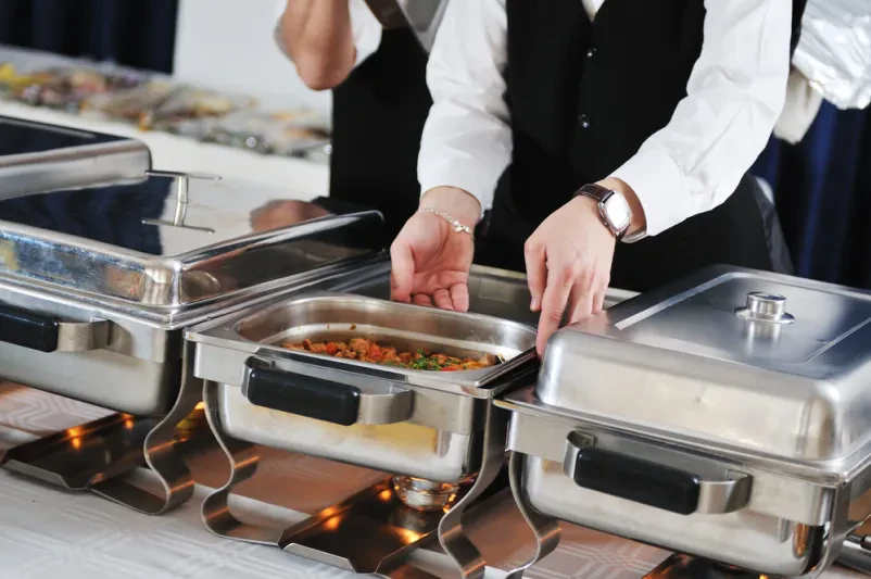 Kuhar v beli uniformi skrbno aranžira izbrane jedi za catering buffet, poudarjajoč kakovost priprave hrane in vizualno privlačnost.