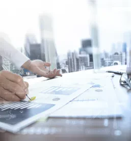 Poslovna miza je polna finančnih dokumentov, pametnega telefona, digitalne tablice in poslovnih grafikonov. V ozadju deluje poslovnež, kar poudarja pomen izdelave analiz in pregledov.