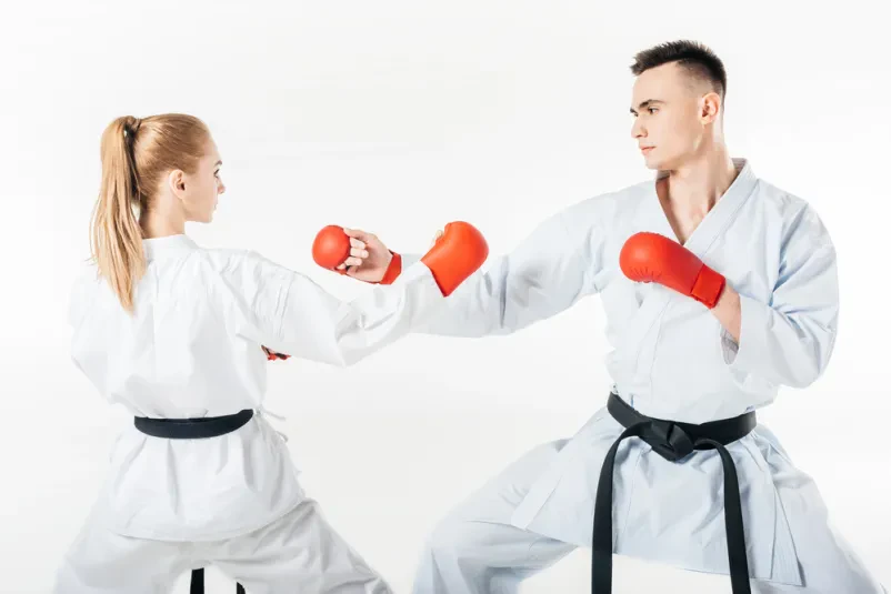 Karate borca v skupnem treningu Moški in ženska karate borec z rdečim in črnim pasom med skupinskim treningom na beli podlagi. Izvajata tehniko bloka in napada v borilnih veščinah.