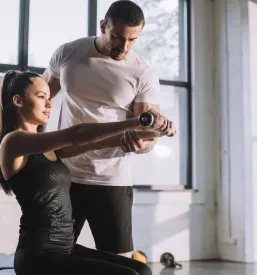 Trener skrbno spremlja in pomaga športnici pri izvedbi vaj z ročnimi utežmi, kar zagotavlja učinkovit in varen trening.