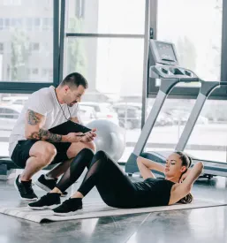 Moški osebni trener s štoparico nadzoruje atletsko žensko, ki na fitnes blazini izvaja vaje za trebuh – osebno trenerstvo v Podravju.