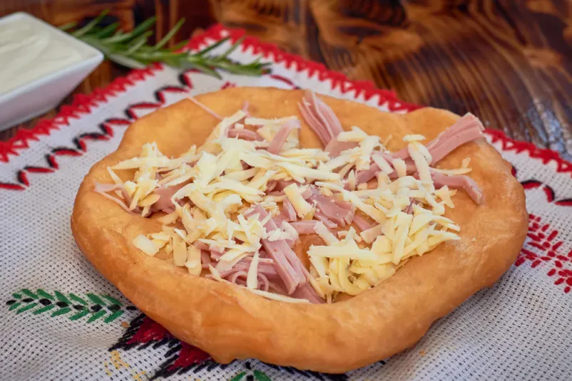 Hrustljav langoš s sirom in slanino Tradicionalni madžarski langoš, ocvrt v olju, obložen z naribanim sirom in hrustljavo slanino, tipična ulična hrana.