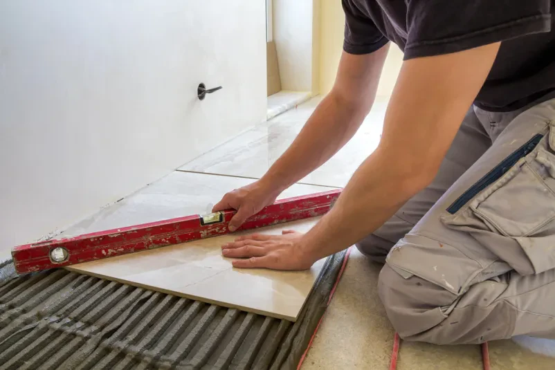 Keramičar s pomočjo ročice namešča keramične ploščice na cementna tla, pod katerimi je rdeč električni sistem talnega gretja. To zagotavlja udobje in kakovostno Polaganje keramike Ajdovščina, Vipava, Nova Gorica.