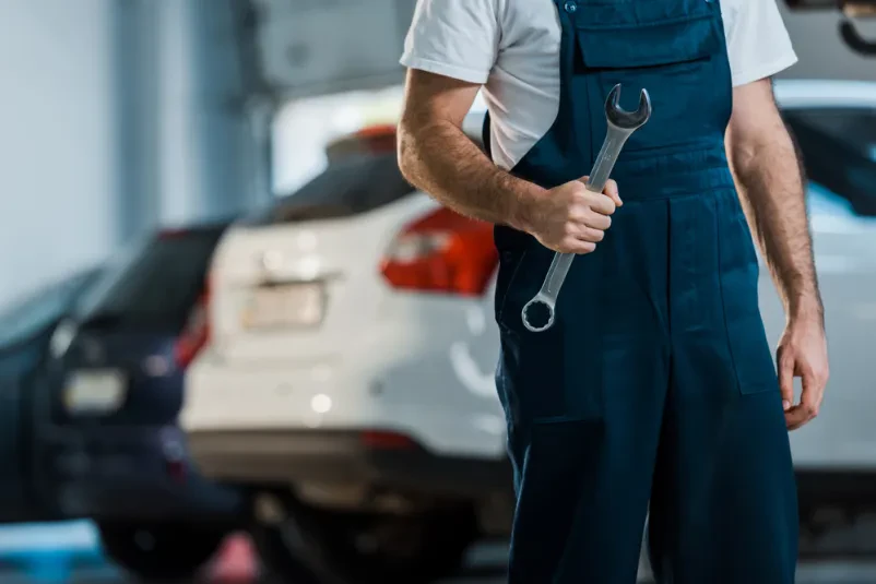 Bližnji posnetek mehanika, ki drži velik viličasti ključ. Prikazuje pripravljenost na delo in natančnost pri avtoservisnih opravilih.