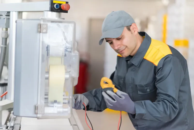 Industrijski električar v delovni obleki in z orodjem v roki predano popravlja industrijski stroj, s poudarkom na natančnosti in strokovnosti pri vzdrževalnih delih.