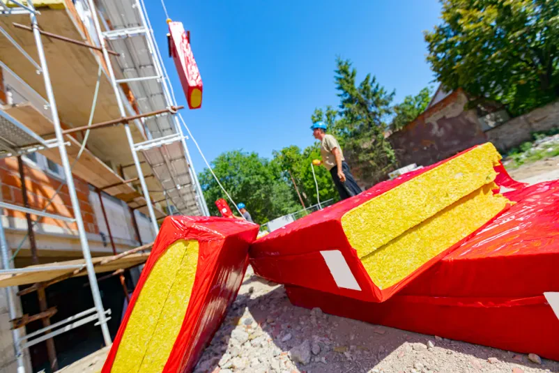 Delavec na gradbišču namešča izolacijo Velik kup toplotnoizolacijskega materiala, ovitega v folijo, na gradbišču. Delavec s pomočjo dvigala namešča en paket kamene volne na kavelj, da ga dvigne na fasado in izolira stanovanjsko stavbo.