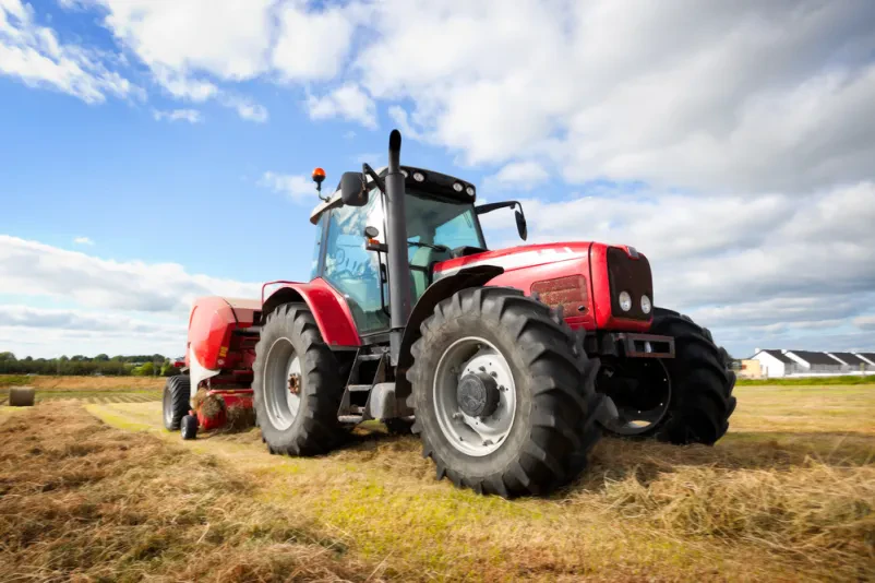 Velik traktor pobira bale sena na zeleni poljani pod modrim nebom, kar je ključno delo pri kmetijski mehanizaciji.