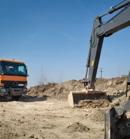 Hidravlični bager v akciji, polni tovornjak z izkopanim materialom, kar kaže na učinkovitost in hitrost ugodnih zemeljskih del v Posavju.