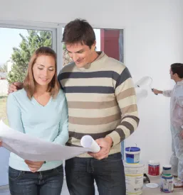 Par si v stanovanju ogleduje načrt za prenovo, v ozadju pa slikopleskar barva steno, kar ponazarja celovit proces adaptacije.