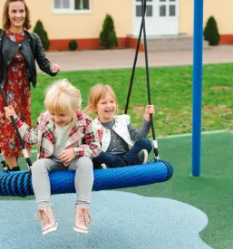 Mlada mati in njeni hčerki se veselo gugajo na sodobni gugalnici v parku, uživajoč v skupni igri in otroškem smehu na prostem.