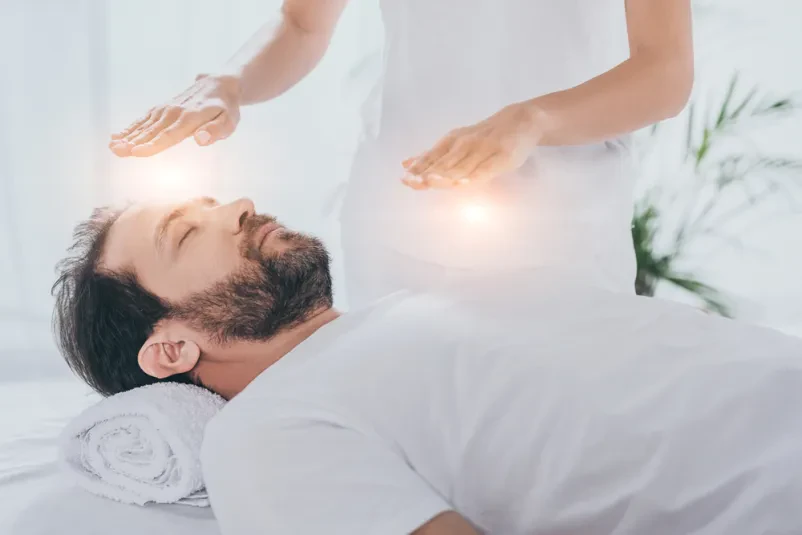 Moški med reiki zdravljenjem Moški z zaprtimi očmi leži sproščen, medtem ko prejema reiki zdravljenje. Fokus je na njegovem notranjem miru.