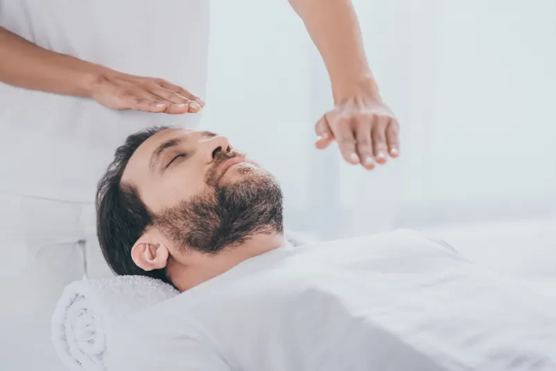 Moški prejema reiki terapijo Spokojen bradati moški leži na masažni mizi, medtem ko terapevt izvaja reiki seanso s položenimi rokami.