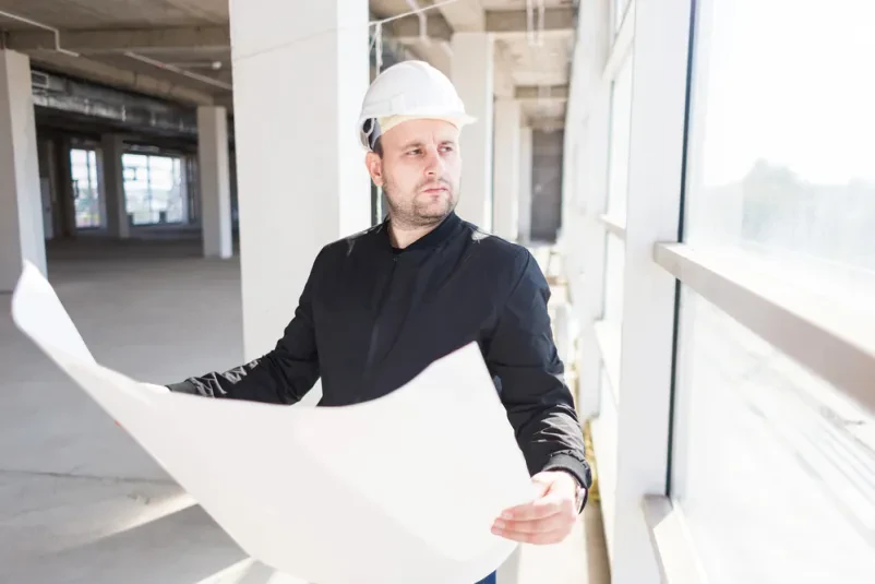 Gradbeni inženir v zaščitni čeladi s projektnimi načrti v roki na gradbišču, ki natančno pregleduje potek del in zagotavlja kakovost.