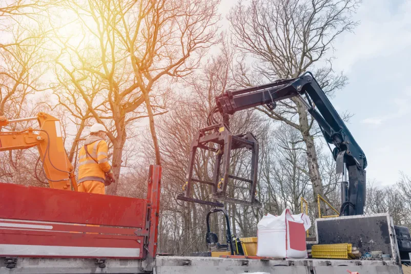 HIAB tovornjak in delavec na gradbišču Delavec z zaščitno čelado stoji ob HIAB tovornjaku z dvigalom, ki je pripravljen za raztovarjanje gradbenega materiala. Poudarek je na varnosti pri delu.