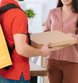 Kurir s termo nahrbtnikom predaja zložene škatle s picami nasmejani poslovni ženski v pisarni, kar kaže na zanesljivo in profesionalno dostavno službo.