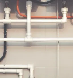 Sistem belih plastičnih vodovodnih cevi z Y-razcepom in črno valovito odtočno cevjo na steni s keramičnimi ploščicami, kar poudarja kvalitetno izvedbo vodovodnih in odtočnih napeljav Savinjska.