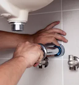 Moški vodovodar v modri delovni obleki montira odtok pod umivalnikom v kopalnici z belimi keramičnimi ploščicami.